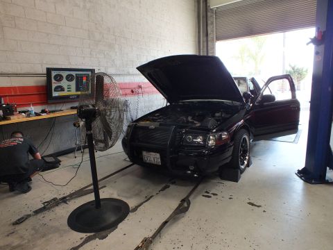 2003 Ford Crown Victoria - black sedan with raised hood and front brush guard sits on a dyno near an open roll-up door, with a fan and a person working in the garage.
