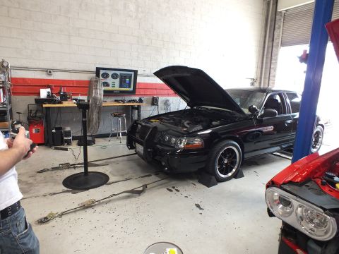 2003 Ford Crown Victoria - black sedan with raised hood sits on a dyno beside a large fan while a person photographs it, with a red car visible in the adjacent garage…