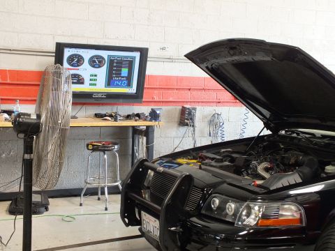 2003 Ford Crown Victoria - open hood and front brush guard of a black car sit beside a large fan and a wall dyno monitor reading 14.0 in a workshop.