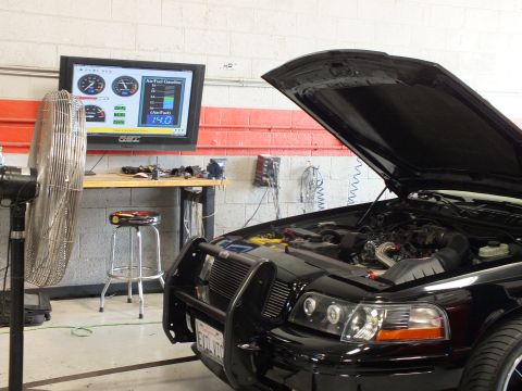 2003 Ford Crown Victoria - raised hood and brush guard of a black car are shown beside a floor fan and a wall-mounted dyno readout monitor in a garage.