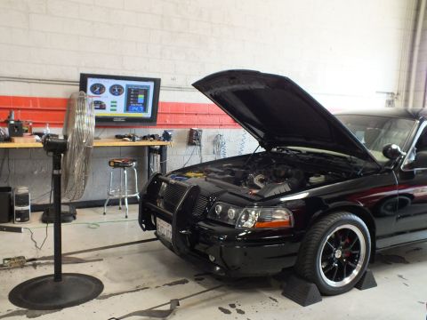 2003 Ford Crown Victoria - black sedan with raised hood and front brush guard sits on a dyno beside a large fan and a wall monitor in a workshop.
