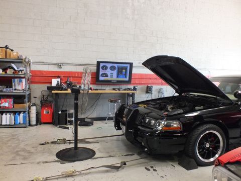 2003 Ford Crown Victoria - black sedan with its hood up and front brush guard sits on a dyno in a workshop lined with shelving, a fan and a wall dyno monitor.