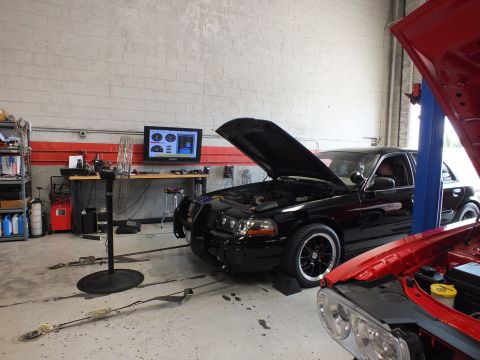 2003 Ford Crown Victoria - black sedan with raised hood and brush guard sits on a dyno beside the open red hood of another car in a busy workshop.