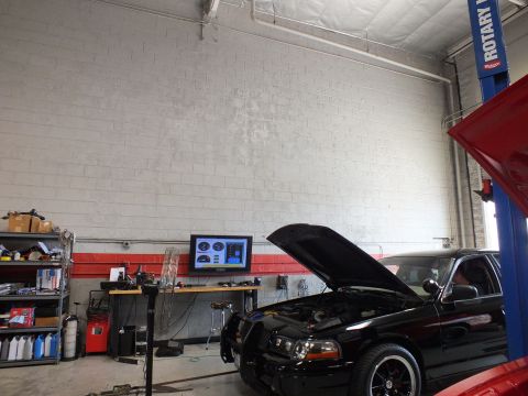 2003 Ford Crown Victoria - black sedan with its hood up sits on a dyno under a two-post lift beside a red car's open hood in a tall white-brick garage.