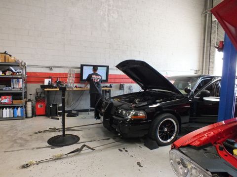 2003 Ford Crown Victoria - black sedan with raised hood and front brush guard sits on a dyno while a man stands at a wall monitor, with a red car hood in the foregrou…
