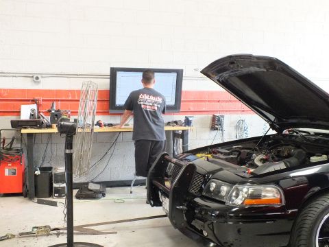 2003 Ford Crown Victoria - front of a black sedan with its hood up sits on a dyno while a man works at a wide wall-mounted monitor in the garage.