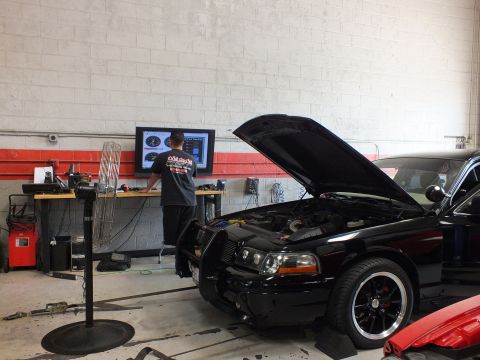 2003 Ford Crown Victoria - black sedan with raised hood and front brush guard sits on a dyno while a man works at a wall dyno monitor in a workshop.
