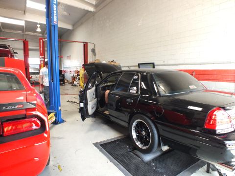 2003 Ford Crown Victoria - black full-size sedan sits on a chassis dyno inside a shop with its driver door open, beside a red Dodge Challenger coupe.