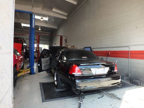2003 Ford Crown Victoria - black full-size sedan rests on the dyno with its driver door open, viewed from the rear three-quarter inside the shop.