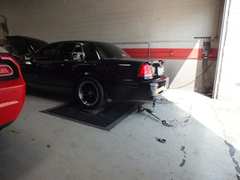 2003 Ford Crown Victoria - rear of a black sedan sits on the dyno pad with its hood up, a red car partly visible at left in the shop.