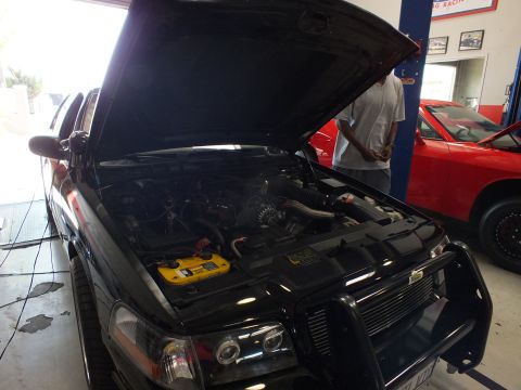 2003 Ford Crown Victoria - black SUV with a brush guard sits with its hood open, exposing the engine bay and a yellow battery box, red car behind.