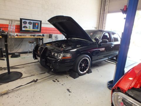 2003 Ford Crown Victoria - black sedan with its hood open sits on the dyno, a colorful gauge results screen mounted on the wall behind it.