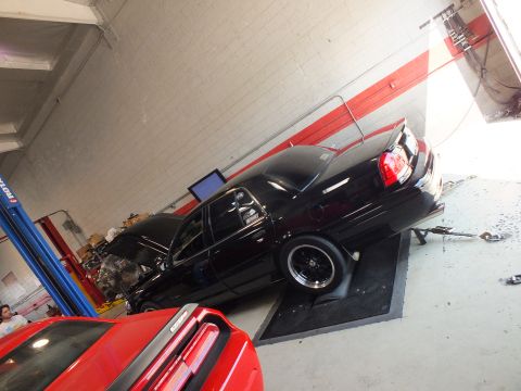 2003 Ford Crown Victoria - canted-angle shot of a black sedan on the dyno pad with a red car's taillight in the foreground inside the shop.