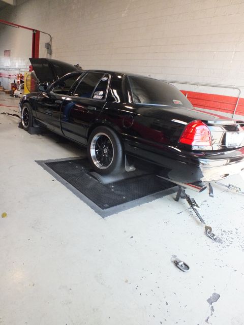 2003 Ford Crown Victoria - black full-size sedan sits on the dyno with its hood raised, shown from the rear three-quarter in the shop.