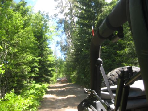 2005 Jeep Wrangler - Two off-road vehicles travel down a dirt road surrounded by dense green trees.