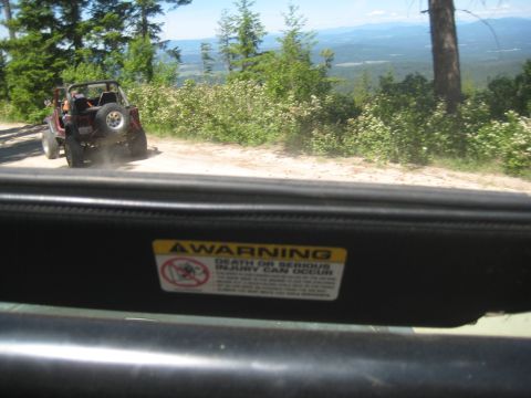 2005 Jeep Wrangler - A Jeep Wrangler kicks up dust on a mountain road, with a vast landscape visible beyond the trees.