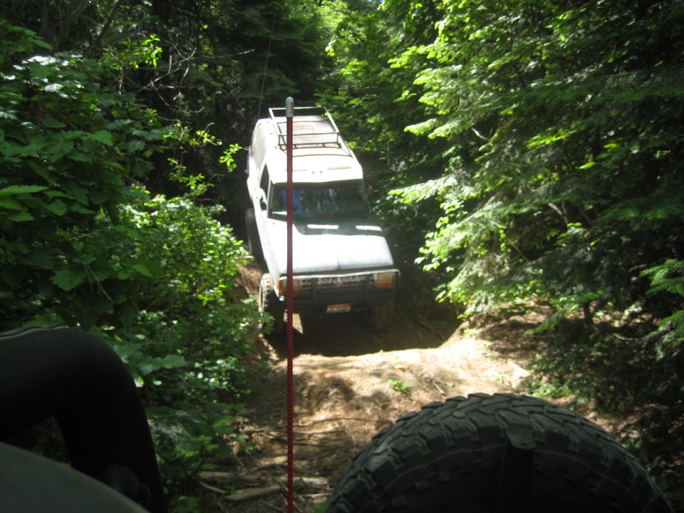 White SUV with a roof rack driving on a narrow dirt trail in a dense forest. Another vehicle's tire is visible in the foreground
