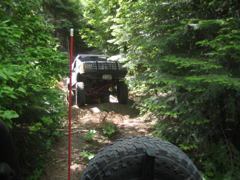 2005 Jeep Wrangler - A lifted Jeep Cherokee XJ navigates a wooded trail, with another vehicle's tire visible in the foreground.