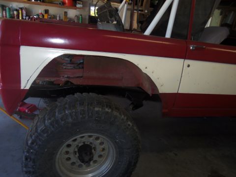 1968 Ford Bronco - The side of a red and white SUV is shown with a large off-road tire in the foreground, undergoing some bodywork.