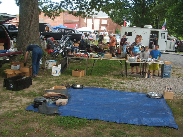 Outdoor swap meet with tables of parts, a motorcycle, and several vehicles in the background