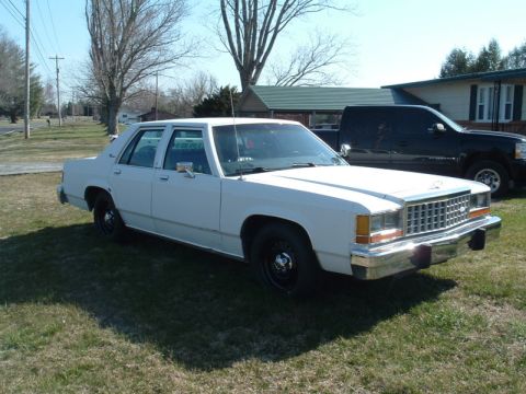 1991 Ford LTD Crown Victoria - A white sedan is parked on a grassy area with a black pickup truck in the background.