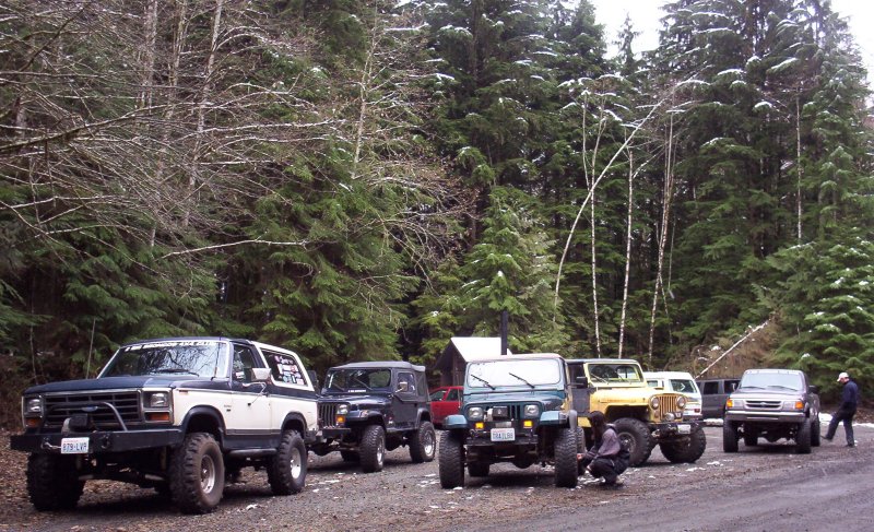 Lineup of lifted 4x4 Broncos and Jeeps parked on a gravel lot in snowy evergreen forest