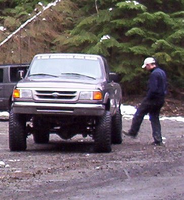 1982 Ford Bronco - A man kicks the tire of a lifted Ford Ranger pickup truck parked on a gravel surface with trees in the background.