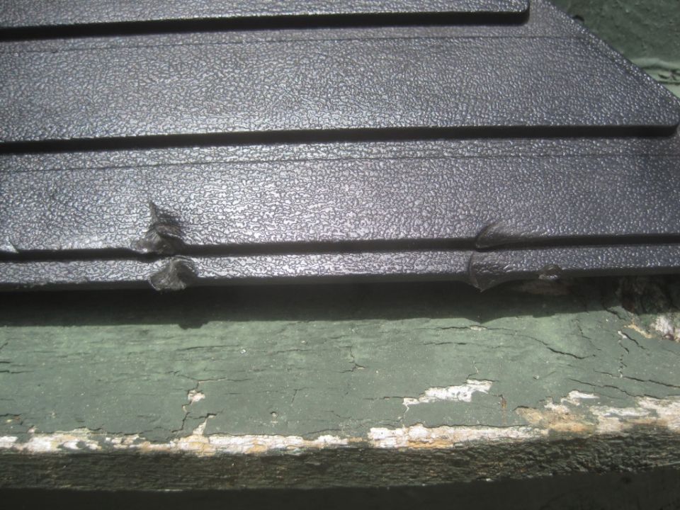Close-up of damaged black plastic trim with horizontal ridges. Green painted wood is below