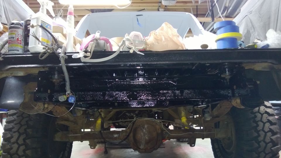 Rear view of a Ford Bronco undergoing work, showing the axle, frame, and suspension components. The bed is visible above, with tools and supplies