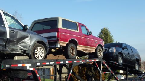 1995 Ford Bronco - A red 1995 Ford Bronco with tan trim is seen on a car carrier alongside other vehicles.