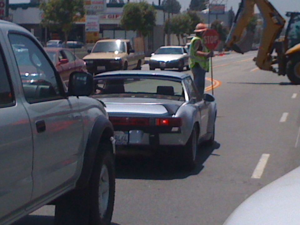Silver sports car in traffic, person holding stop sign next to it. License plate reads 7976