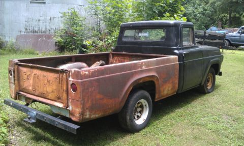1957 Ford F-100 - A weathered 1957 Ford F-100 pickup truck sits in a grassy area next to a building, with another vehicle visible in the background.