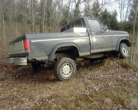 1985 Ford F-150 - Three-quarter rear view of a dark gray/black square-body Ford regular-cab longbed 4x4 flexing its suspension on an off-camber leaf-covered…