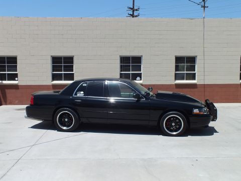 2003 Ford Crown Victoria - black full-size sedan on black wide wheels is parked in profile on concrete outside a gray-and-brick building.