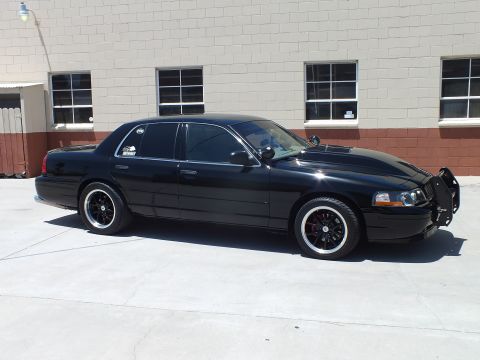 2003 Ford Crown Victoria - black full-size sedan fitted with a front push bar sits in front three-quarter view on concrete outside a gray building.
