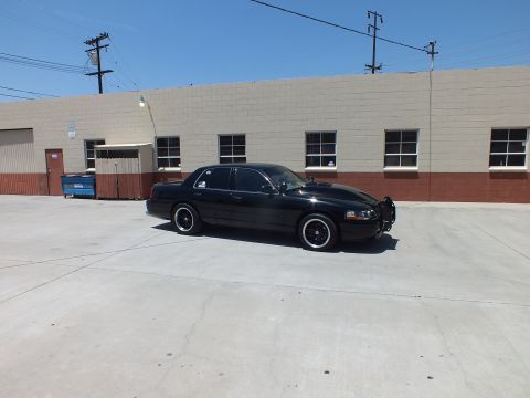2003 Ford Crown Victoria - black full-size sedan is parked at an angle in an open concrete lot beside a blue dumpster and a long building.