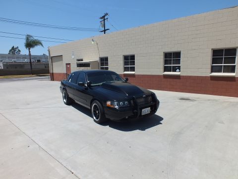 2003 Ford Crown Victoria - black sedan with a front push bar and halo headlights sits in front three-quarter view on concrete by a brick building.