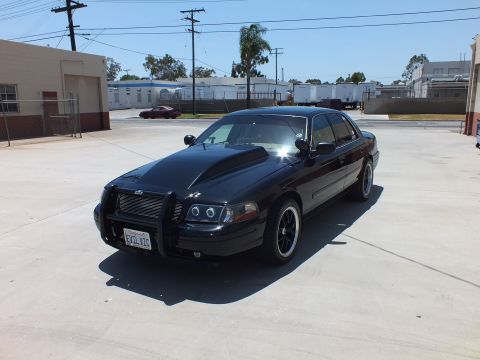 2003 Ford Crown Victoria - black full-size sedan with a front push bar and halo headlights sits in a sunny concrete lot near trees and buildings.