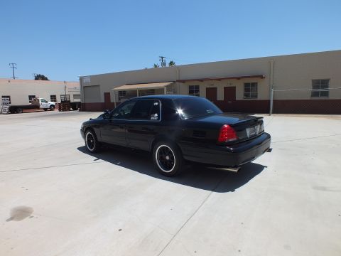 2003 Ford Crown Victoria - black full-size sedan on dark wheels sits in rear three-quarter view in an open concrete lot before a tan warehouse.