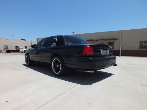 2003 Ford Crown Victoria - black full-size sedan with dual exhaust tips sits in rear three-quarter view on concrete near a fence and warehouse.