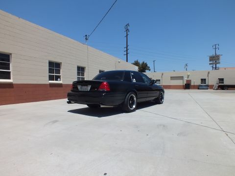 2003 Ford Crown Victoria - black full-size sedan sits in rear three-quarter view in an open concrete lot beneath a clear blue sky by gray buildings.