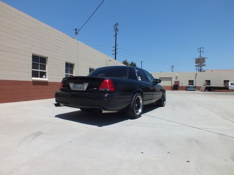 2003 Ford Crown Victoria - black full-size sedan with dual exhaust sits in rear three-quarter view in a wide concrete lot under a blue sky.