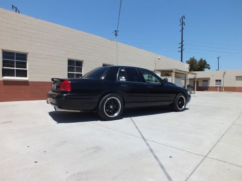 2003 Ford Crown Victoria - black full-size sedan on dark wheels sits in rear-side view on concrete in front of a long tan-and-brick building.