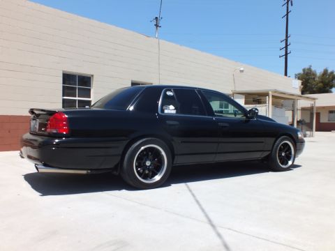 2003 Ford Crown Victoria - black full-size sedan sits in rear three-quarter view on concrete before a tan building with a covered entry.