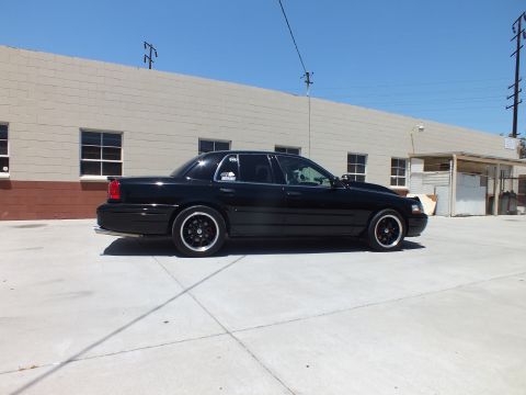 2003 Ford Crown Victoria - black full-size sedan on dark wheels is parked in profile on concrete in front of a long tan building under blue sky.