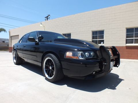 2003 Ford Crown Victoria - black full-size sedan with a front push bar and halo headlights sits in front three-quarter view on concrete by a building.