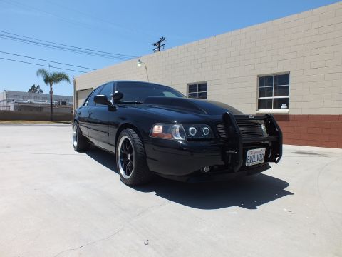 2003 Ford Crown Victoria - black full-size sedan with a push bar and halo lights sits outdoors, its plate reading EVIL VIC, by a gray building.