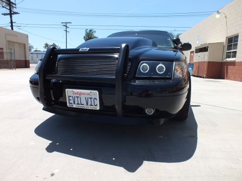 2003 Ford Crown Victoria - head-on close-up of a black sedan's push bar and halo headlights, with a California plate reading EVIL VIC and a Crown Victoria frame.