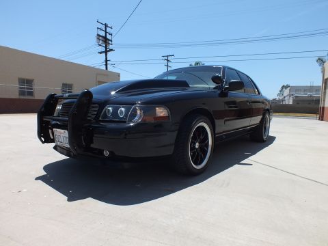 2003 Ford Crown Victoria - blacked-out Ford Crown Victoria with a front push bar and dark wheels sits angled in a sunny industrial lot.