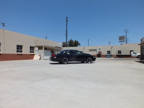 2003 Ford Crown Victoria - black sedan sits alone in a broad concrete lot fronted by tan warehouse buildings under clear sky.